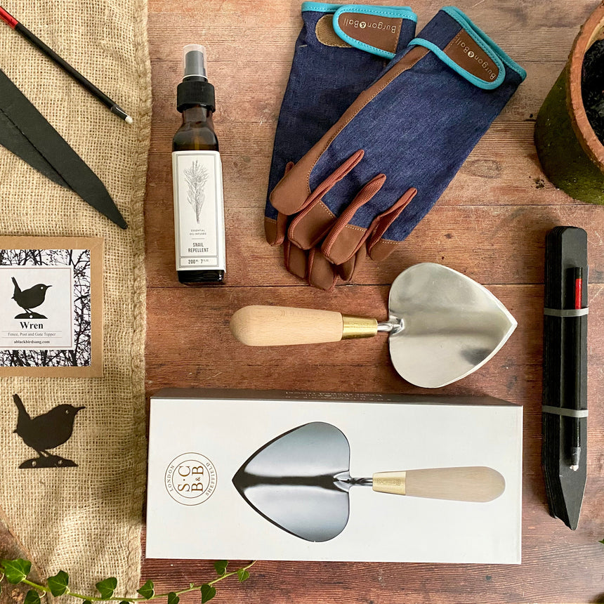 Flat lay of potting Shed gardener's gift box with heart shaped stainless steel trowel, powder coated wren silhoutte garden decoration, blackboard plant markers with pencil, gardening gloves, tool maintenance oil spray in glass bottle