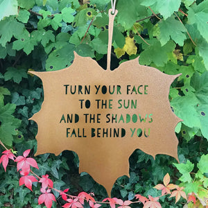 A gold-colored metal leaf-shaped sign with a quote 'Turn your face to the sun, and the shadows fall behind you' hung from a chain, with a background of green leaves.