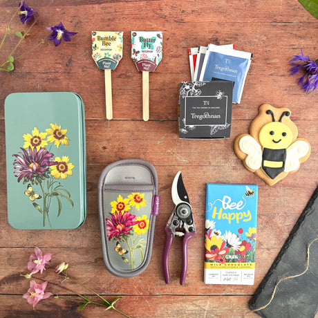 Happy Gardener Gift Box With Secatuers in pouch and tin, bee iced biscuit, selection of black teas, Chocolate bar and two pollinator seed pops with coir and seeds ready to plant. Shown on a dark wooden flatlay background with flowers and twine