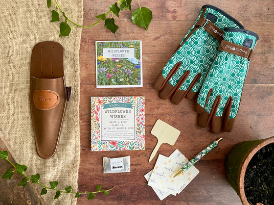 Flatlay with gardening gloves with green art deco pattern, brown leather secatuer pouch, and packet of wildflower wishes with seeds seed paper and a wooden label. Shown on a wood background with ivy, plant pot, a pen and hessian